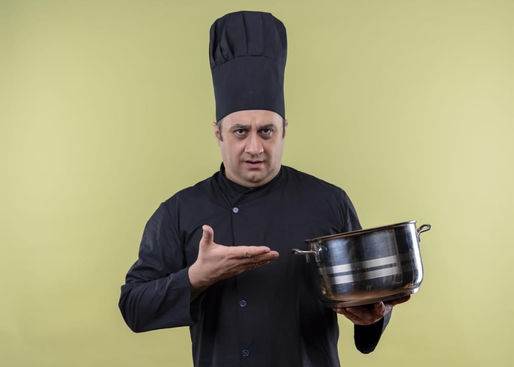 Male-chef-cook-wearing-black-uniform-and-cook-hat-demonstrating-saucepan-with-arm-looking-confused-standing-over-green-background-scaled-1.jpg