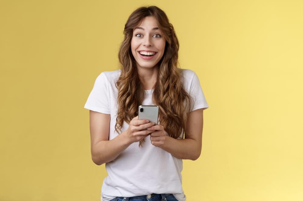 Extremely-happy-enthusiastic-surprised-joyful-lovely-feminine-girl-receive-pleasant-touching-message-online-hold-smartphone-smiling-broadly-look-camera-joyful-amused-yellow-background-scaled-1.jpg